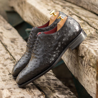Black ostrich leather whole-cut oxford shoes with red lining and wooden shoe trees on a rustic wooden surface.